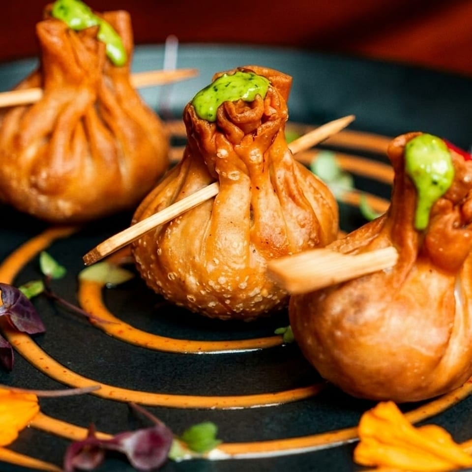 Three golden-fried money bag dumplings with green sauce and skewers on a black plate with orange swirls.