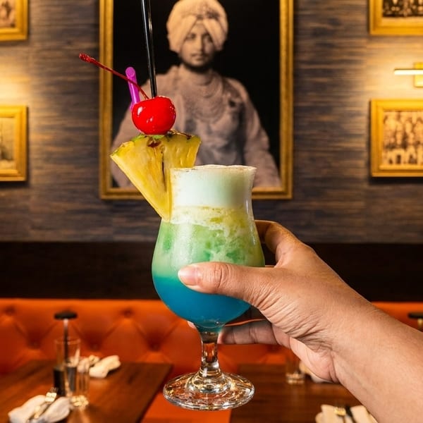 Hand holding a vibrant blue and green cocktail with pineapple and cherry garnish in a restaurant.
