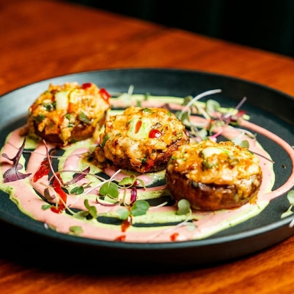 Three stuffed mushrooms with melted cheese, green and pink sauces, and microgreens on a black plate.