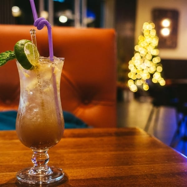 A cocktail with lime and mint on a wooden table in a dimly lit restaurant with festive lights.