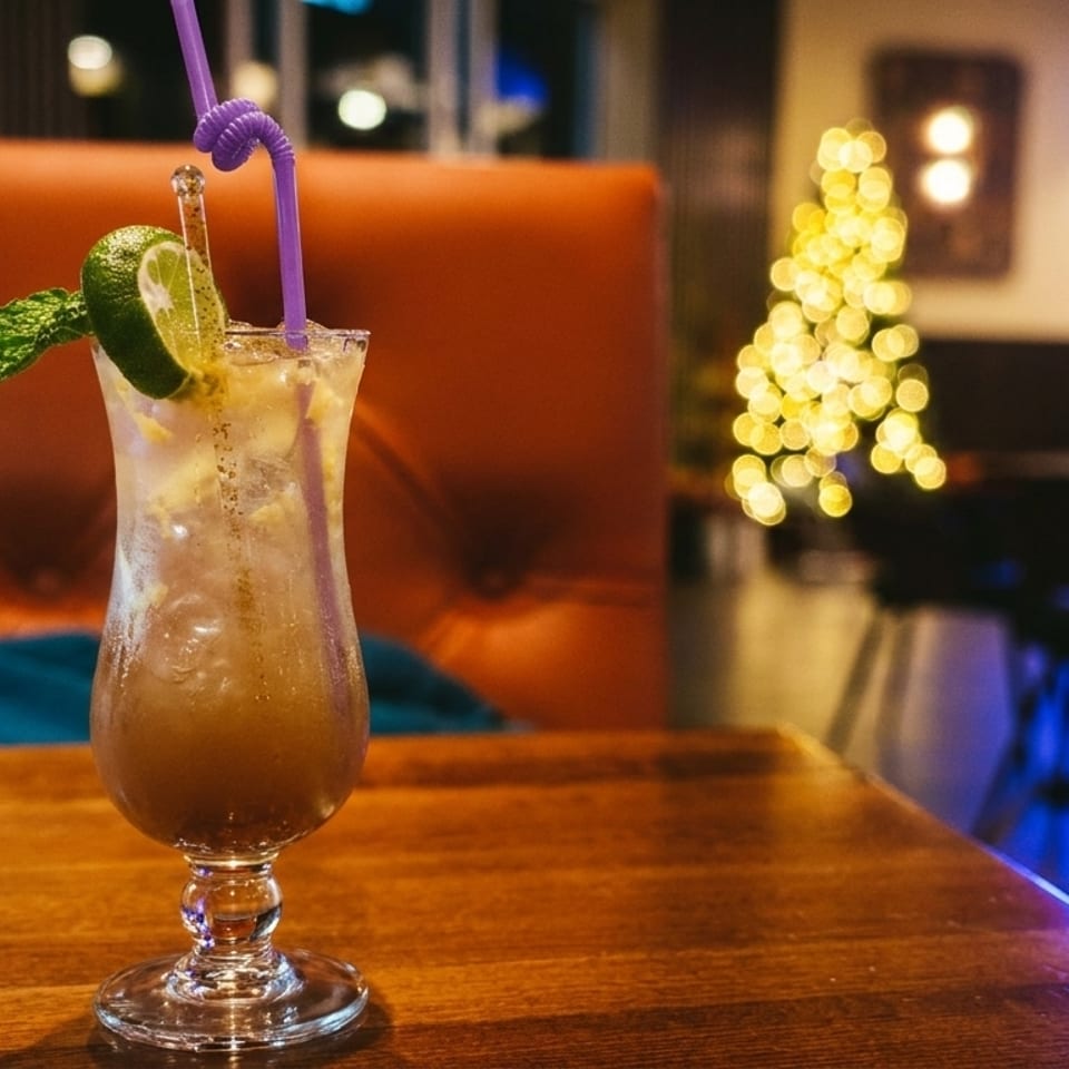 A cocktail with lime and mint on a wooden table in a dimly lit restaurant with festive lights.