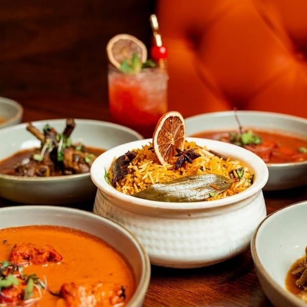 A table laden with various Indian dishes, including biryani, curries, and cocktails, against an orange background.