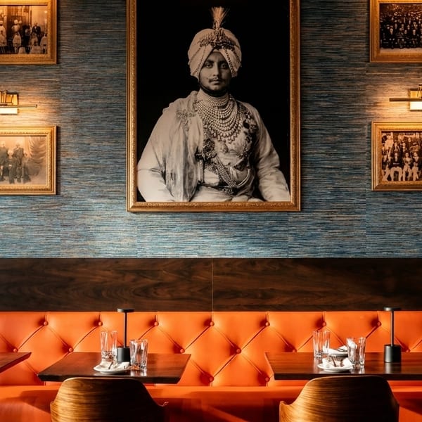 Restaurant interior with orange booths, dark wood tables, and framed portraits on a blue textured wall.